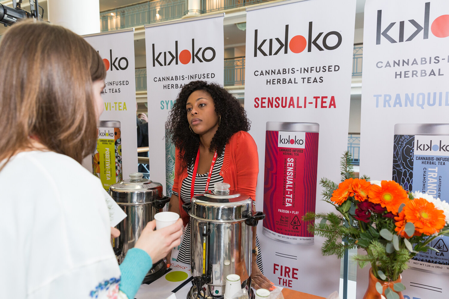 Woman in an orange cardigan pouring tea from a tea urn at a Kikoko booth.