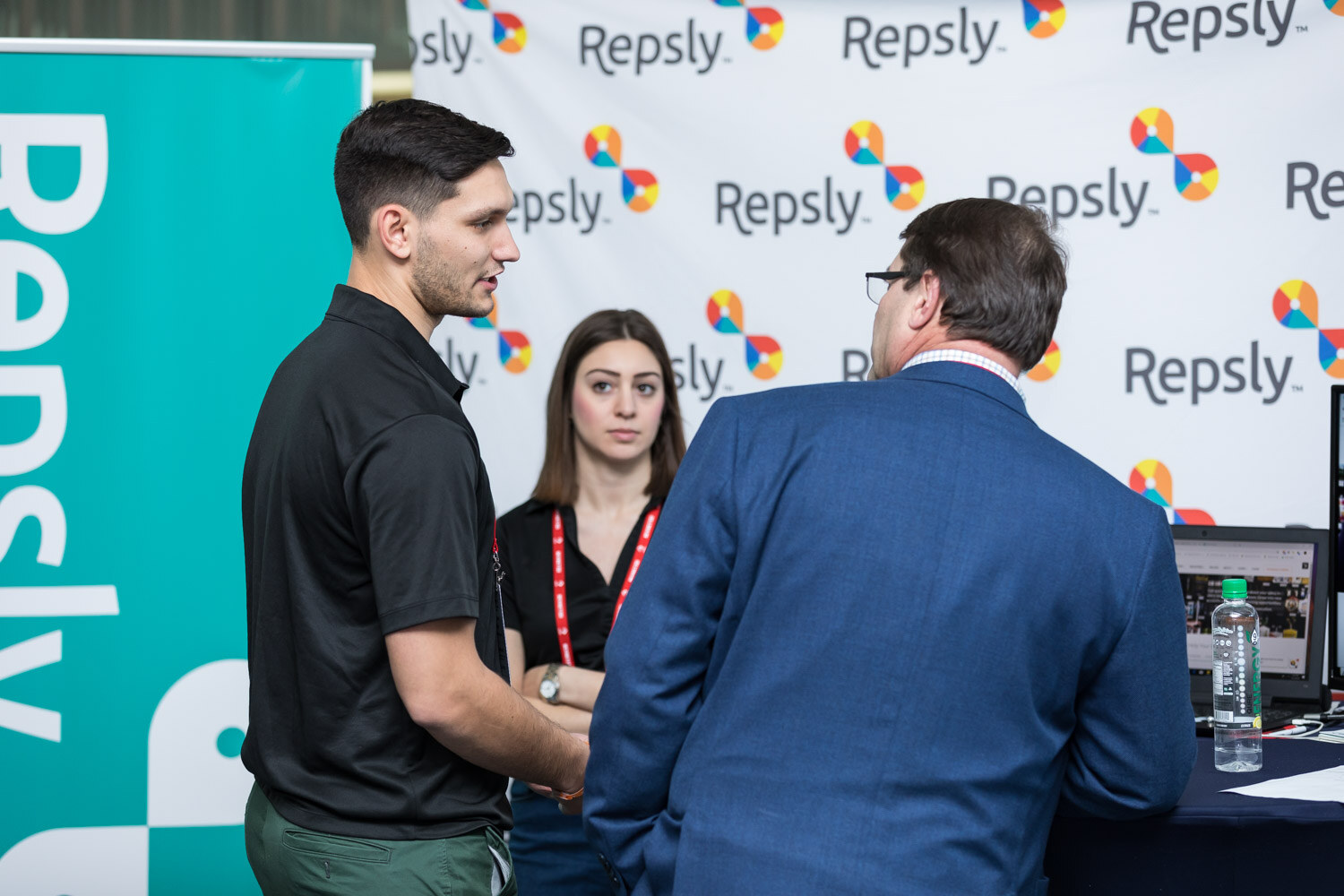 Three people at a booth with a wall of Repsly logos behind them.