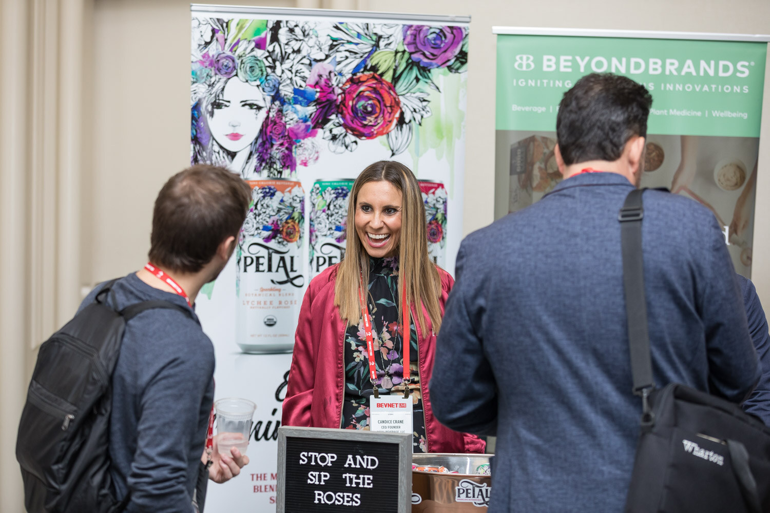 Smiling woman behind a table with Petal and Beyond Brands banners and a sign reading Stop and Sip the Roses.