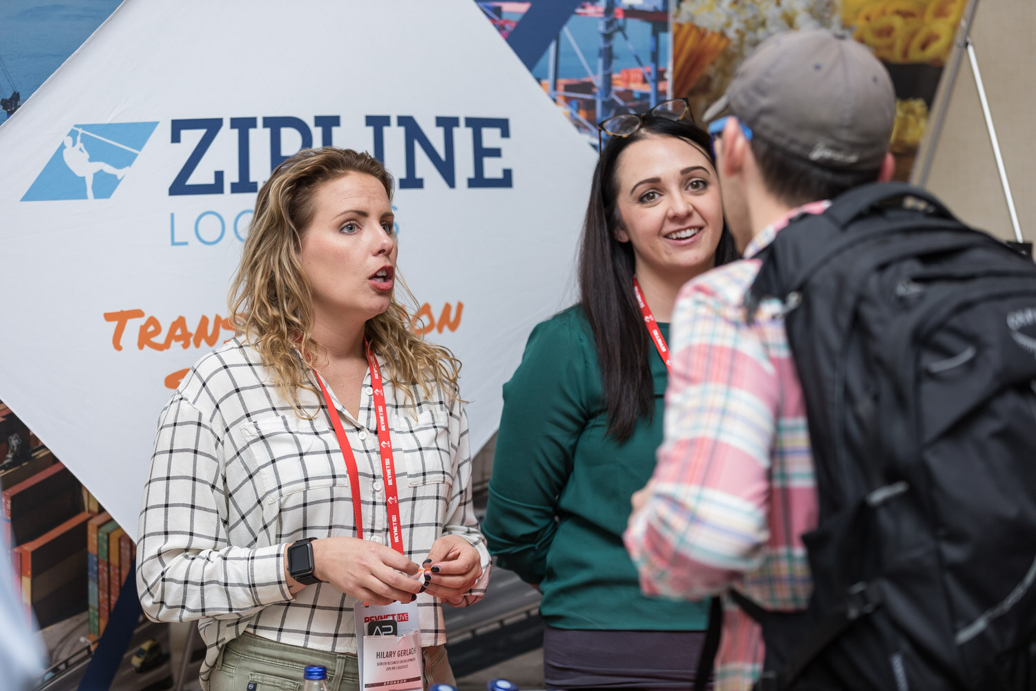 Two women talking with a man in a backpack in front of a Zipline Logistics banner.
