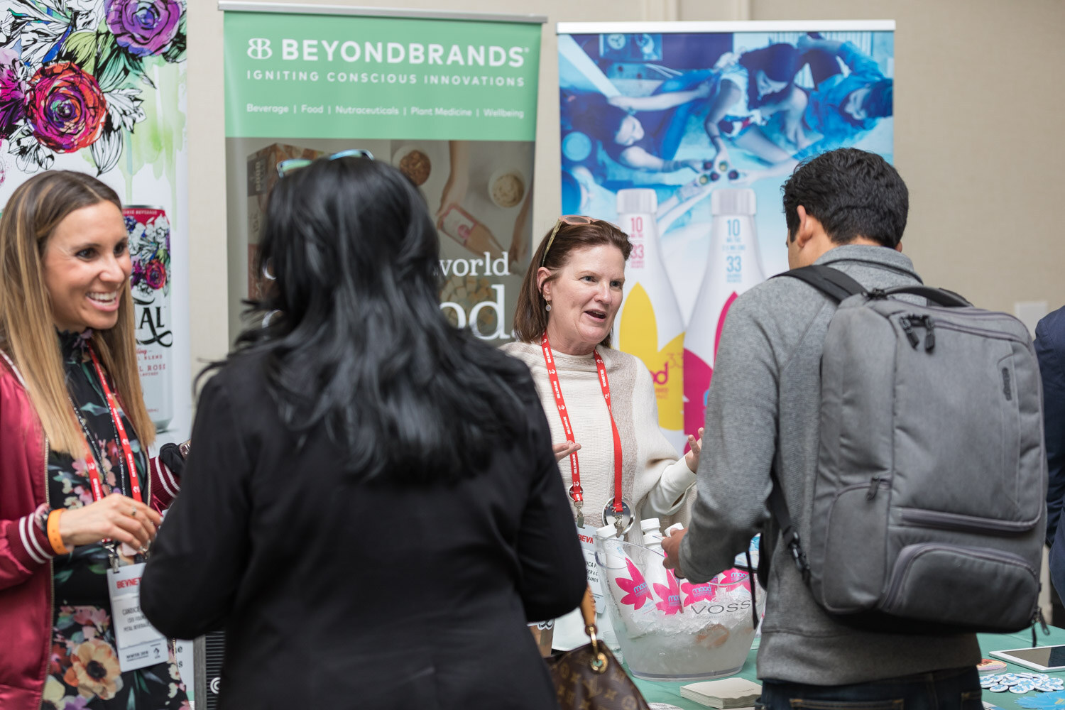 Smiling women at a booth with Beyond Brands banners and a backpacked attendee in front.