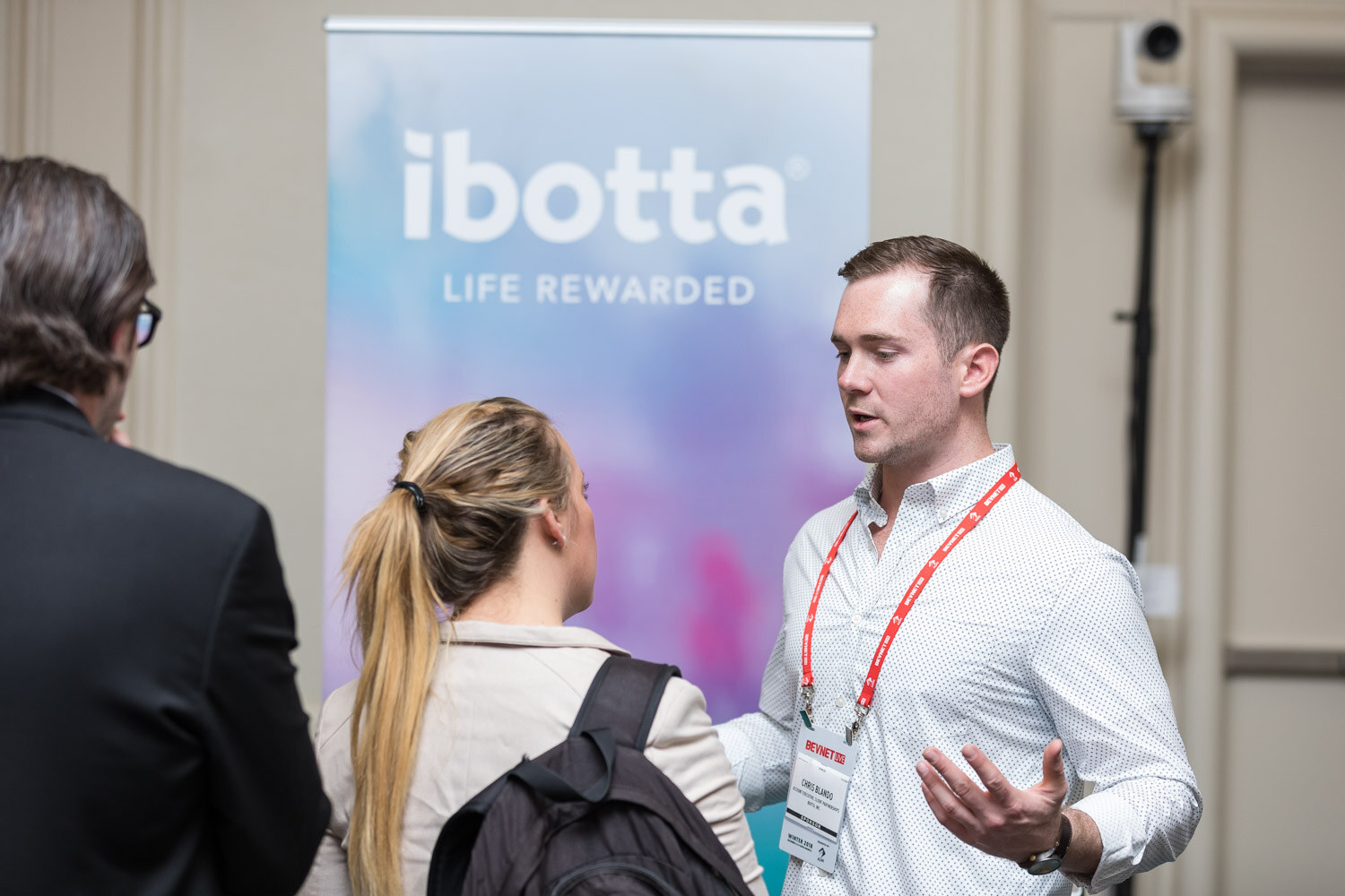 Man in a white shirt gesturing in front of an Ibotta banner with a woman in a backpack listening.