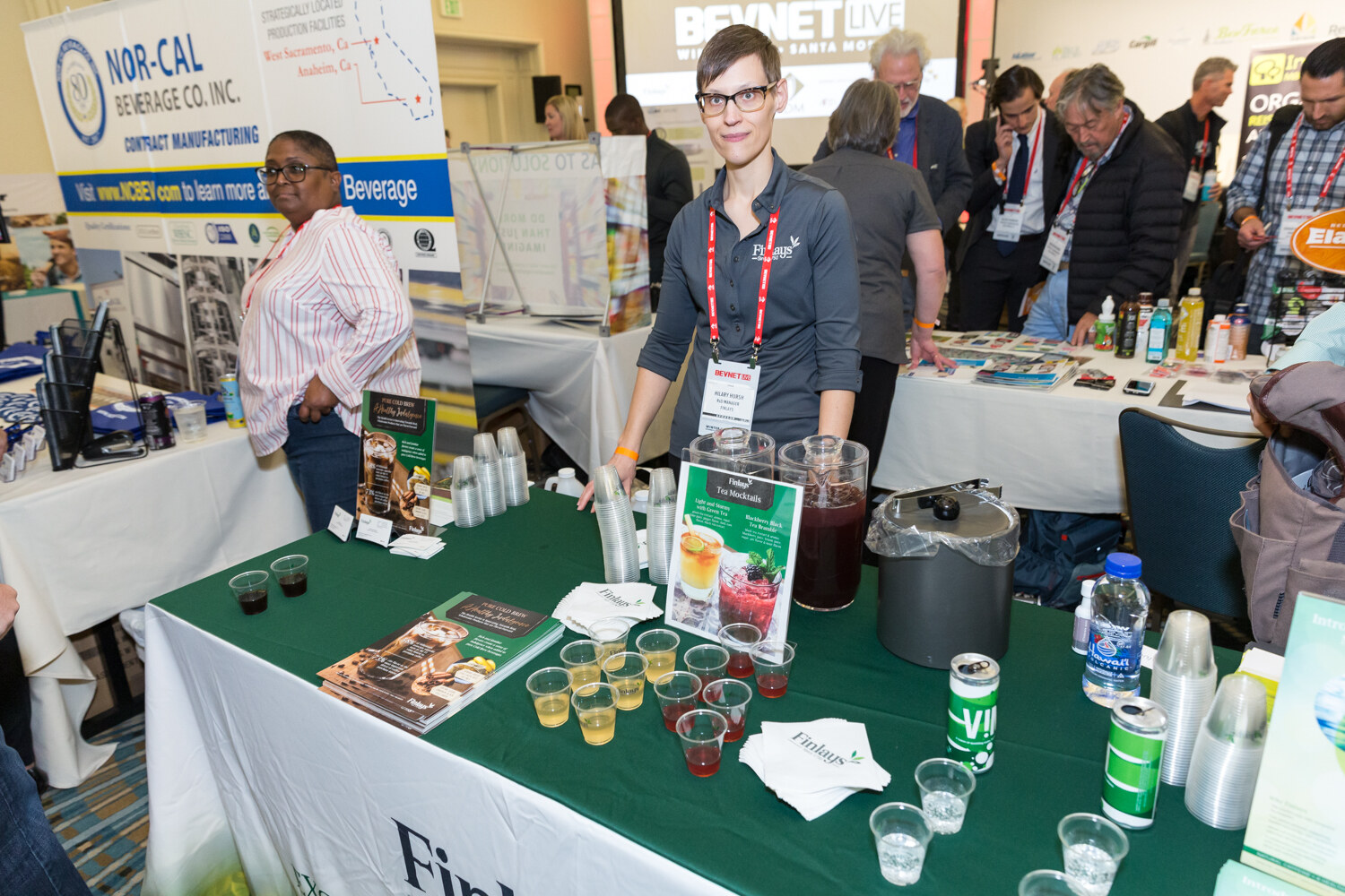 Woman behind a green draped table with sample cups in front of a Nor-Cal banner.