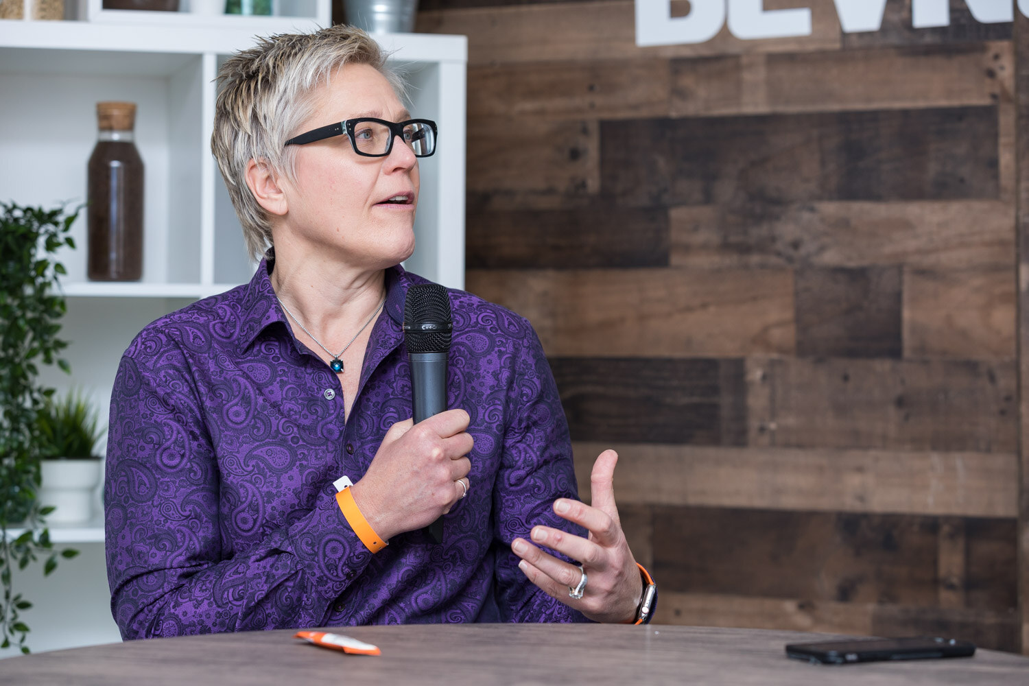 Woman in a purple patterned shirt holding a microphone at a studio table.