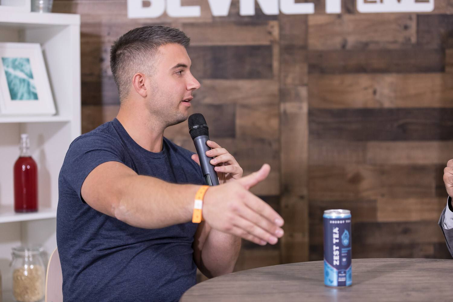 Man in a navy Zest Tea shirt gesturing at a studio table with a can in front of him.