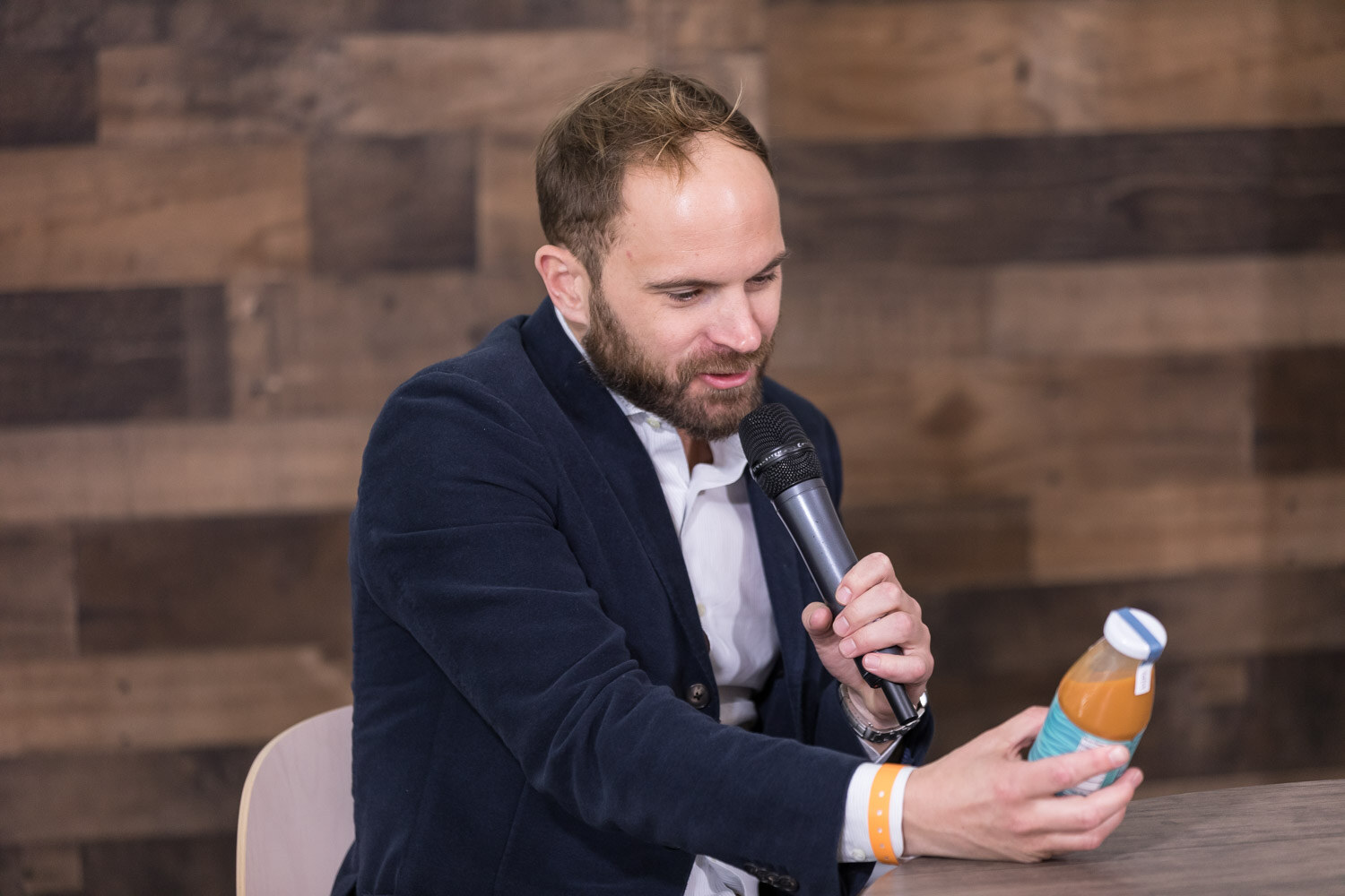 Bearded man in a navy blazer holding a bottle at a livestream studio table.