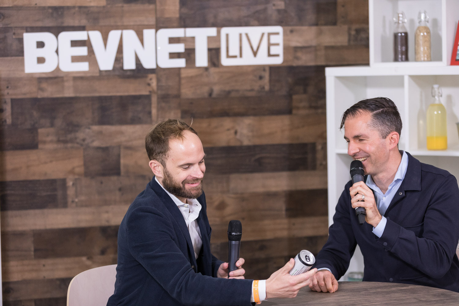 Two men leaning toward each other across a studio table, one passing a can.