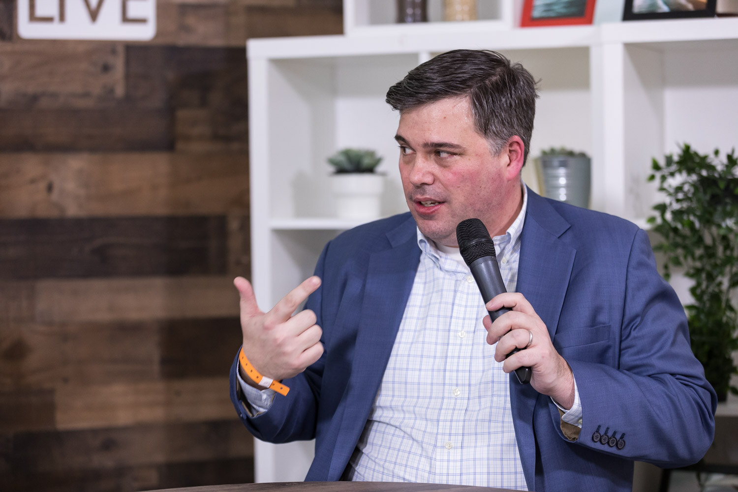 Man in a blue blazer holding a microphone with shelves and a wood wall behind him.