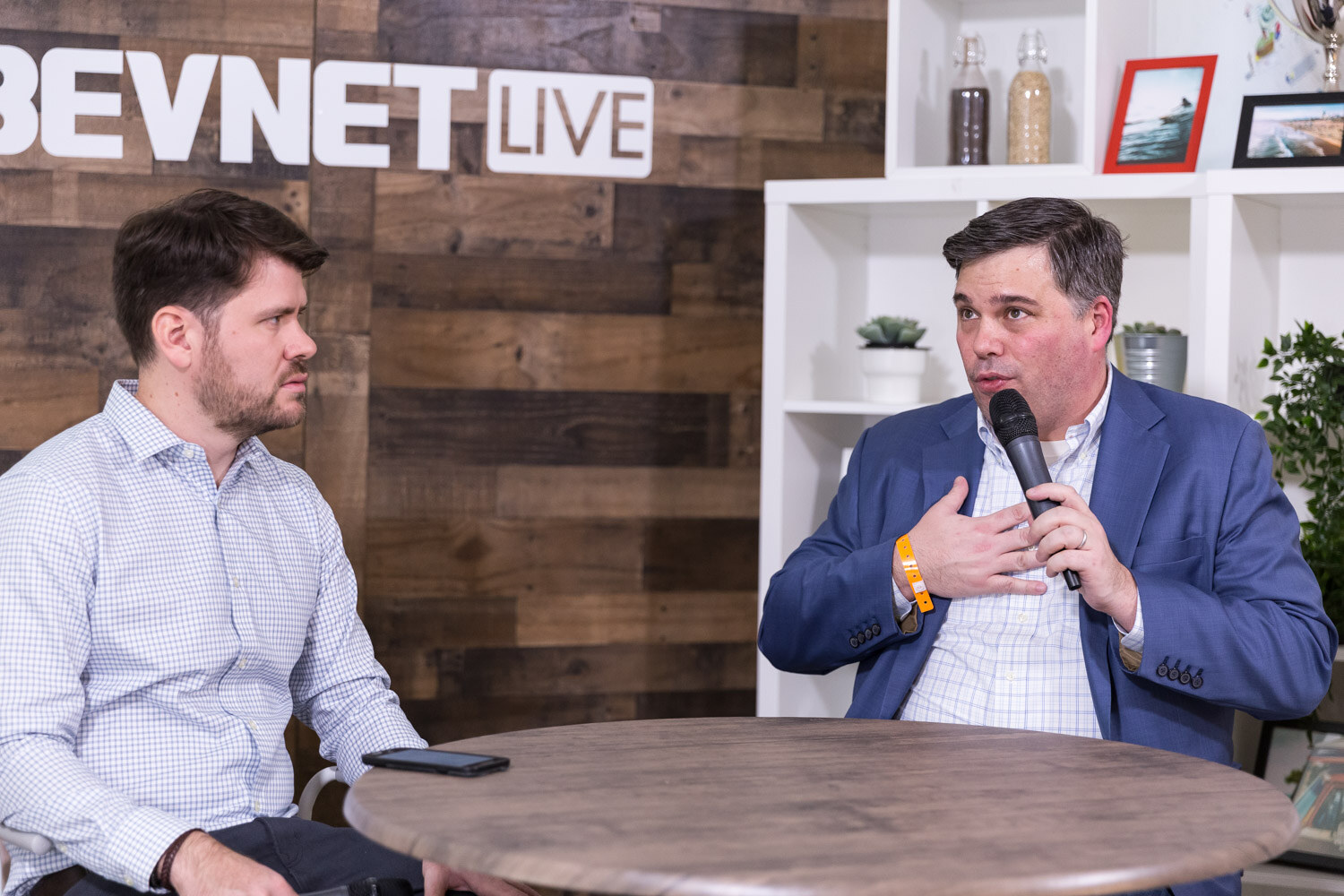 Two men seated at a round table in a livestream studio, one speaking into a microphone.