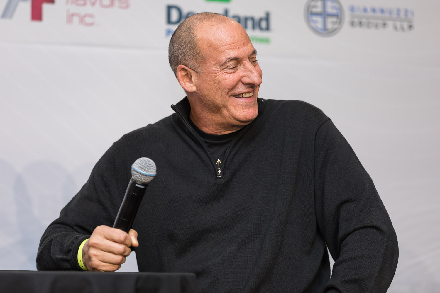 Bald man in a black sweater laughing on stage beside a microphone.