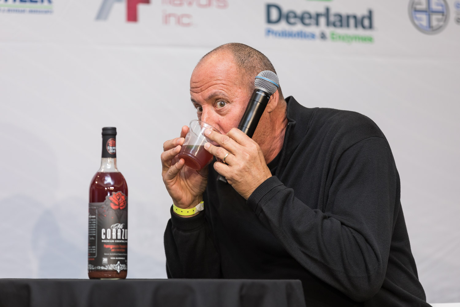 Bald man in a black sweater drinking from a cup with a bottle beside him.