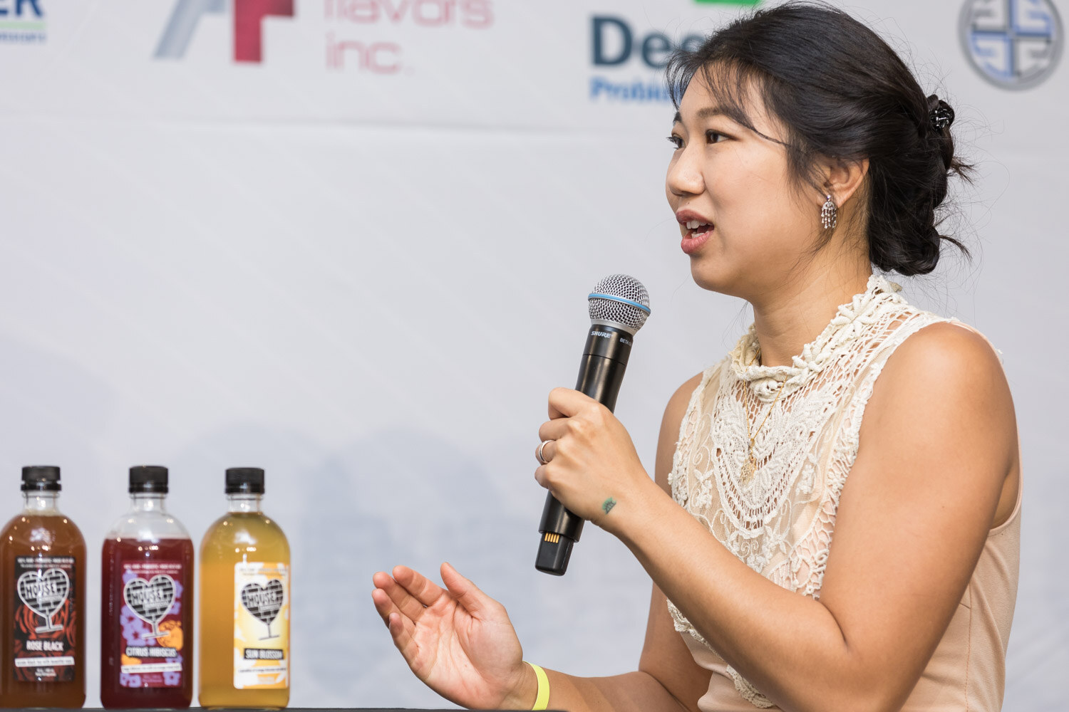 Woman in a cream top holding a microphone beside three bottles of kombucha.