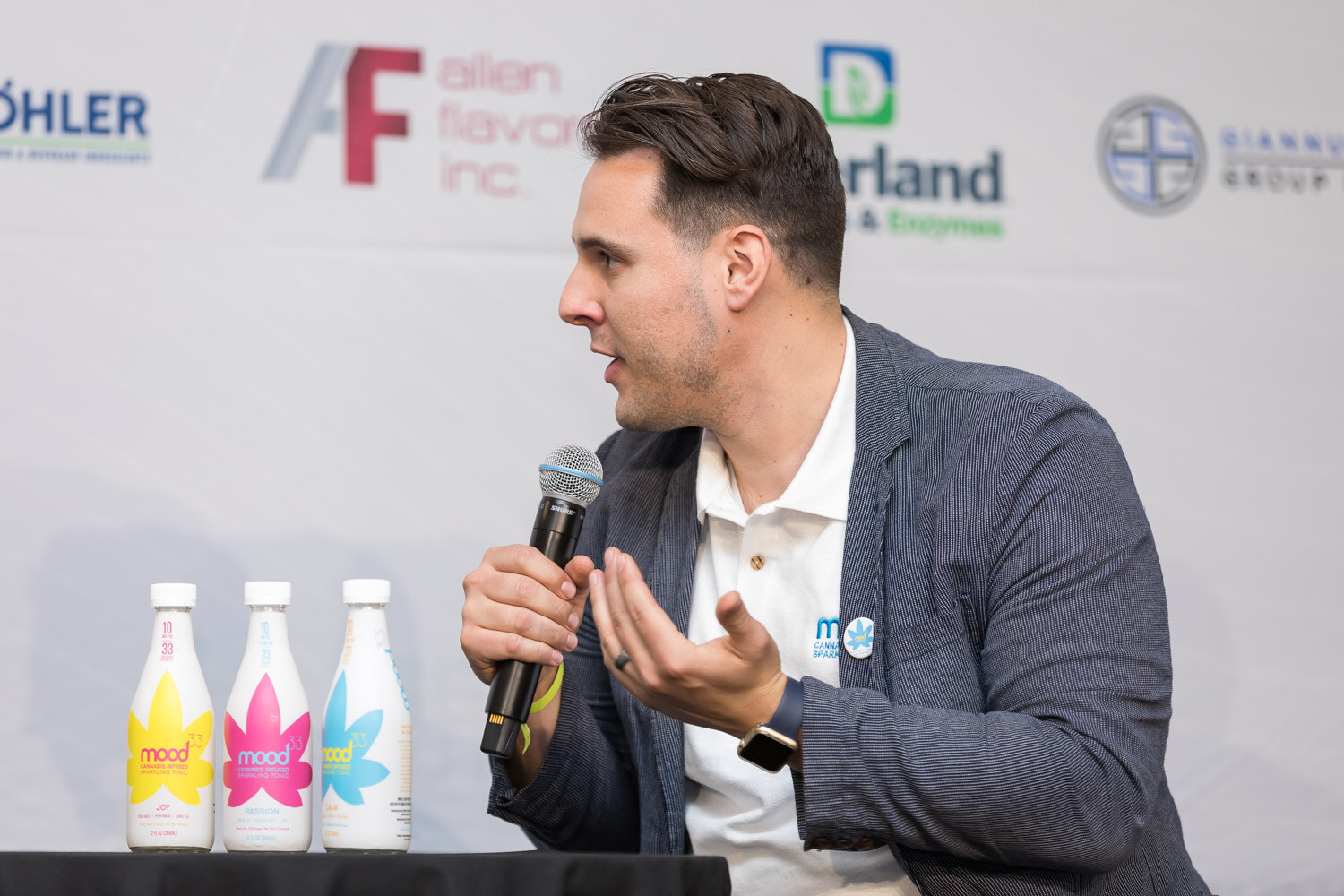 Man in a navy blazer and white shirt talking with three bottles on the table in front of him.