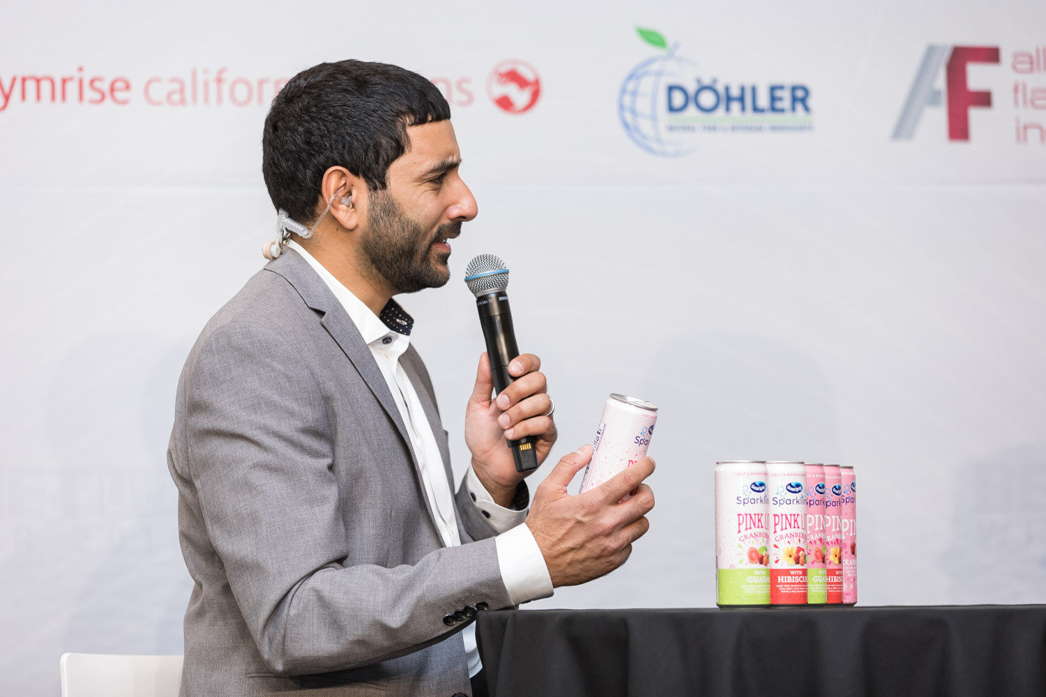 Man in a gray blazer holding a microphone and a pink can beside a stack of cans.