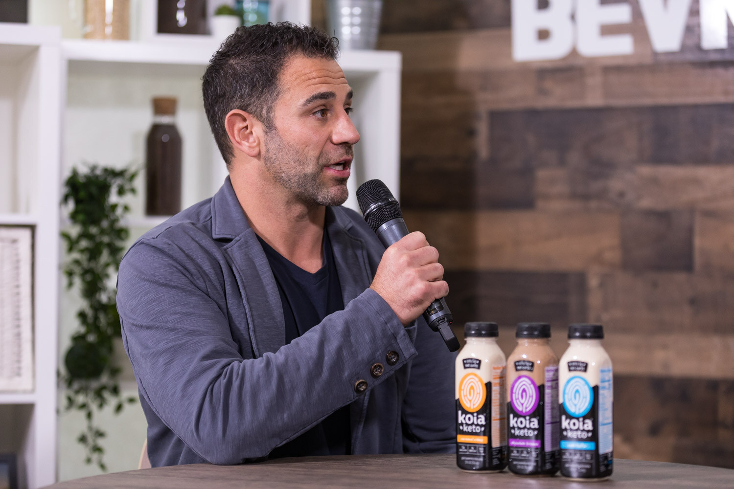 Man in a gray jacket speaking into a microphone with three Koia bottles on the table.