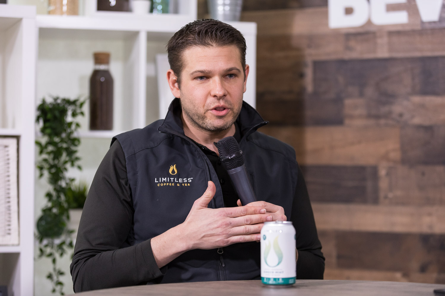 Man in a black vest speaking at a studio table with a white can in front of him.