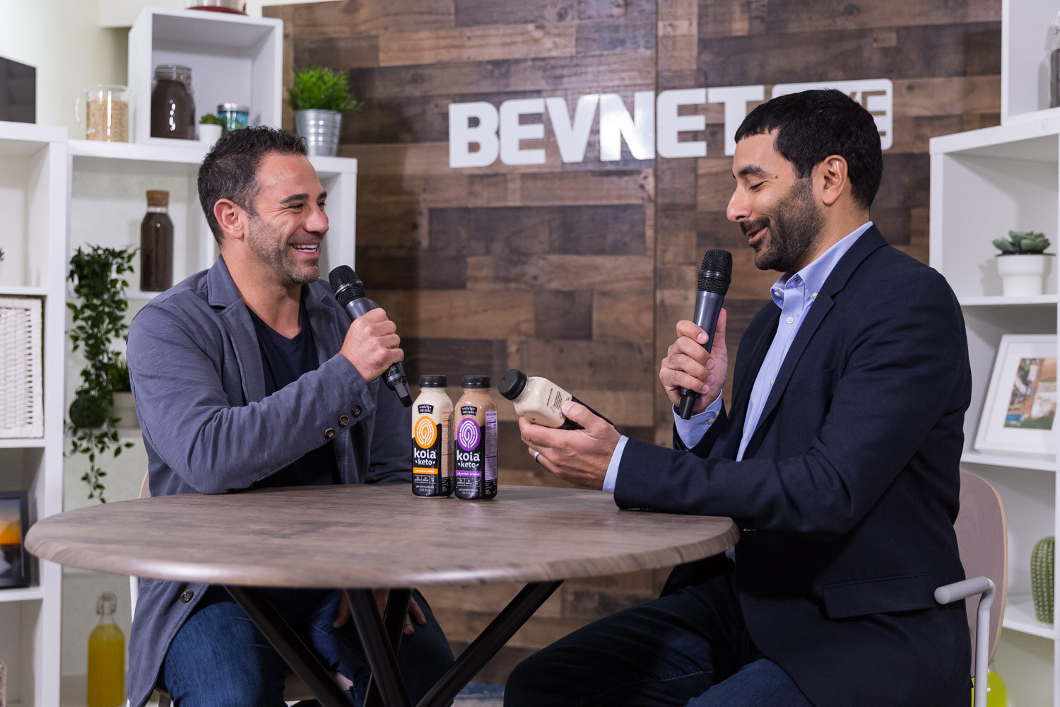 Two men at a studio table holding a bottle and microphones.
