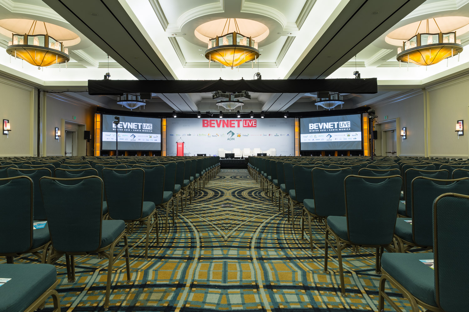 Wide view of an empty ballroom with rows of teal chairs facing a stage and large BevNET Live screens.