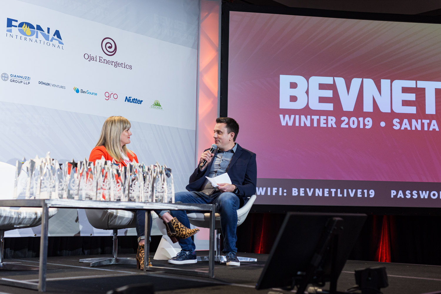 A woman in an orange top and a man holding a microphone sit on stage next to a long table covered in glass award trophies, with a BevNET Winter 2019 screen behind them.