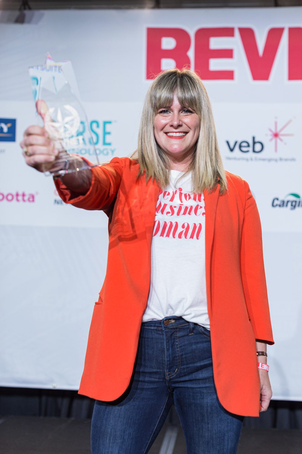 A smiling woman with bangs in an orange blazer and a Serious Business Woman shirt holds a glass award toward the camera.