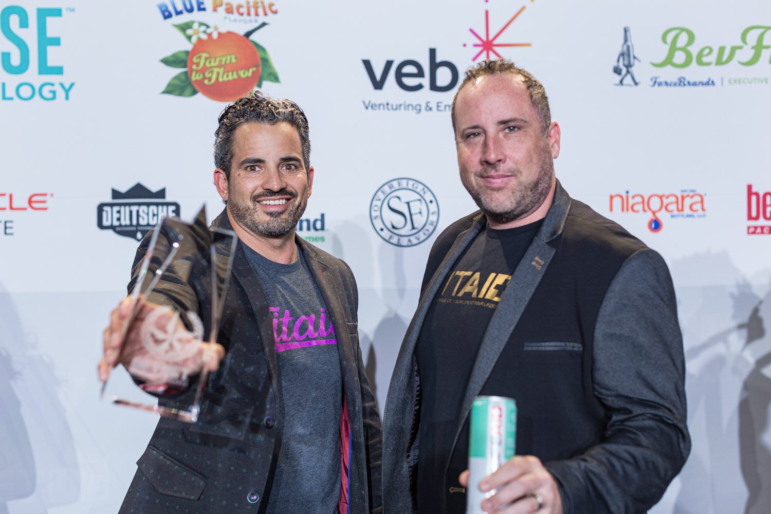 Two men in dark jackets stand in front of a veb and BevForce backdrop, one holding a glass award and the other a can.