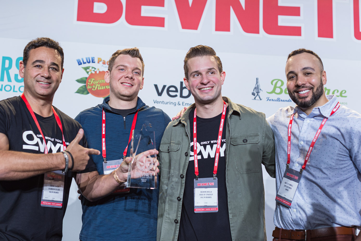 Four men with conference lanyards stand together in front of the sponsor wall, two wearing OWYN shirts and one holding a glass award.
