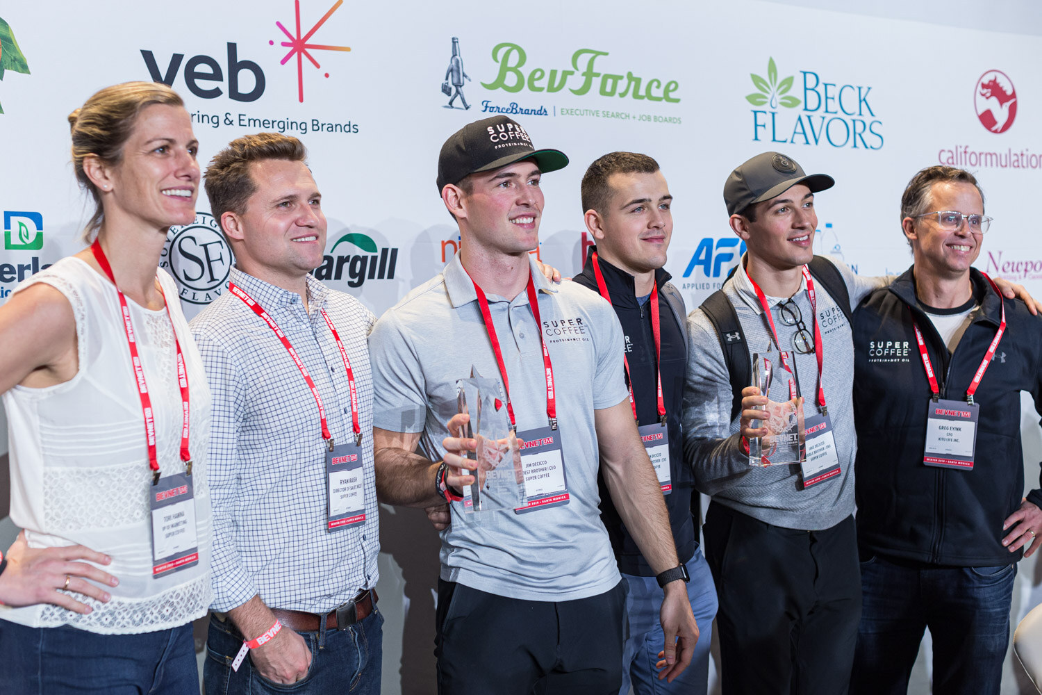 Six people with lanyards, including two in caps, pose in a row in front of the BevForce and veb sponsor wall.