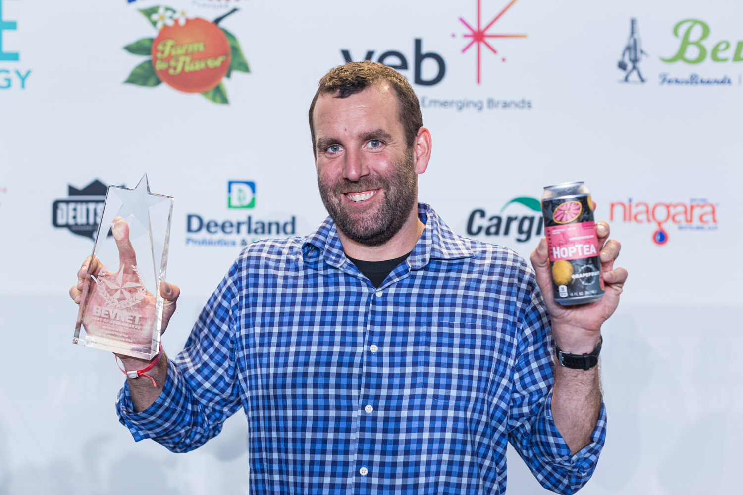 A man in a blue gingham shirt holds a star-shaped glass award in one hand and a HopTea can in the other.