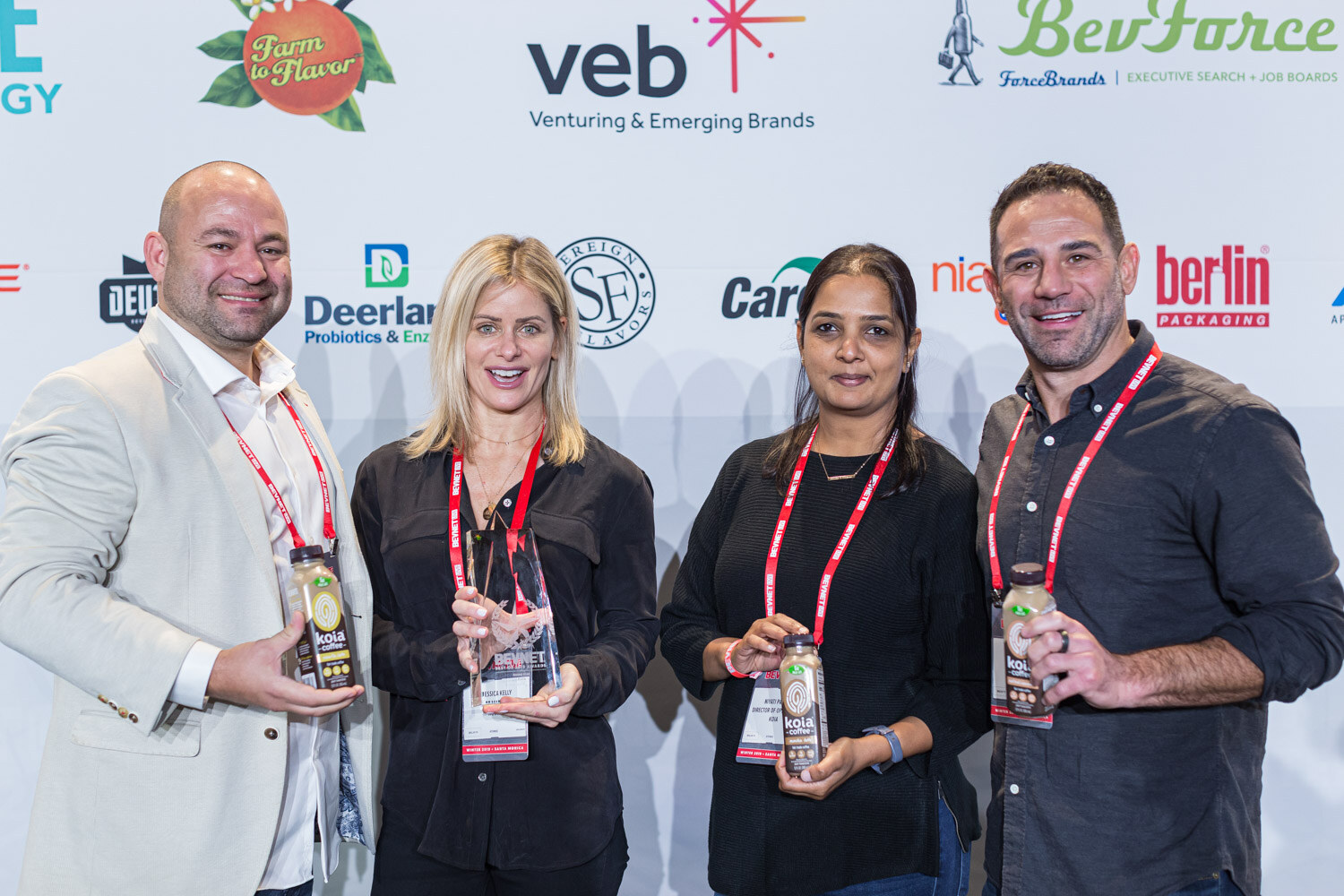 Two men and two women with red lanyards hold bottles and cans while standing in front of the sponsor wall.
