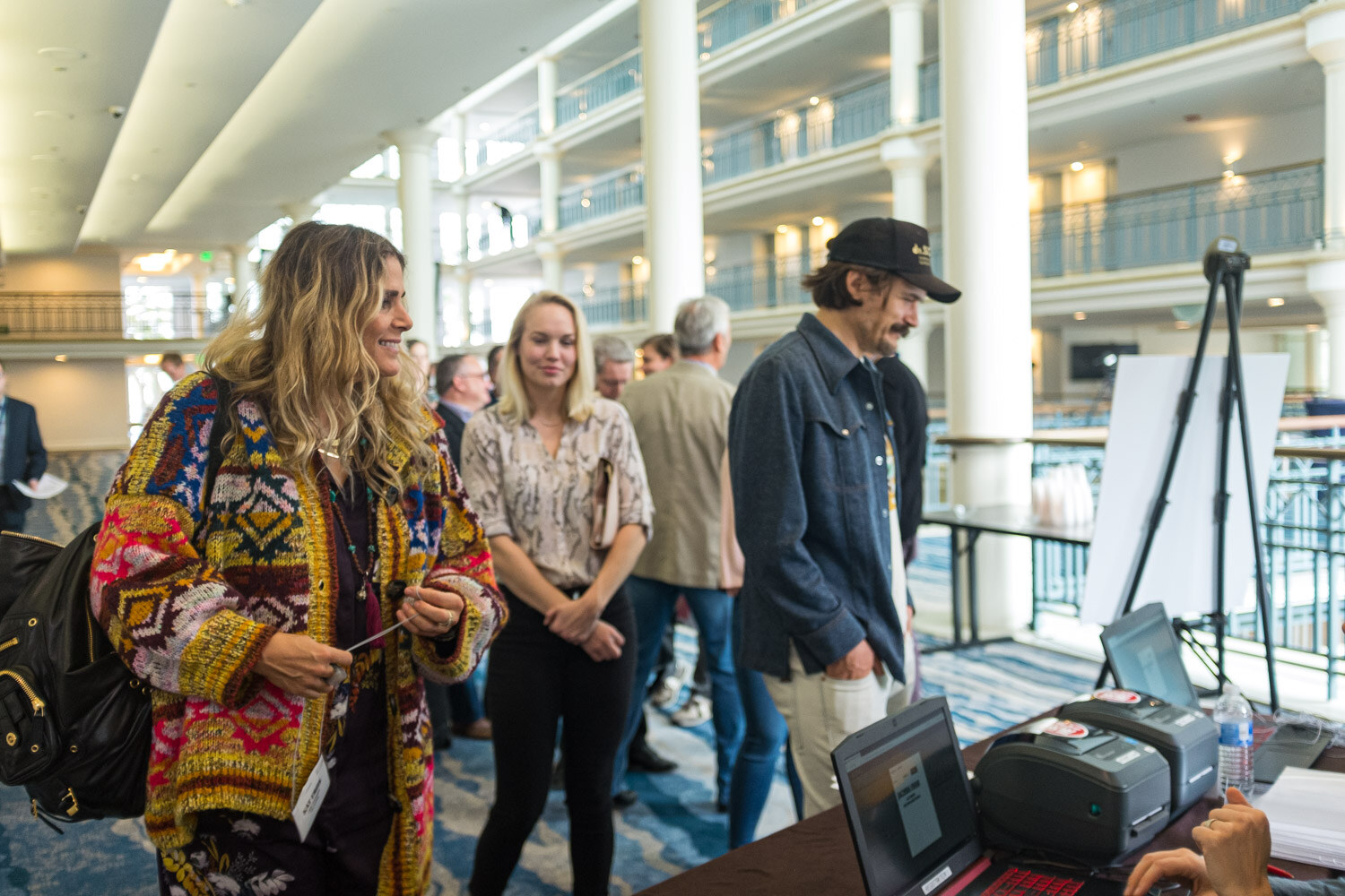 People line up beside a registration table with laptops and a label printer in the multi-story atrium.