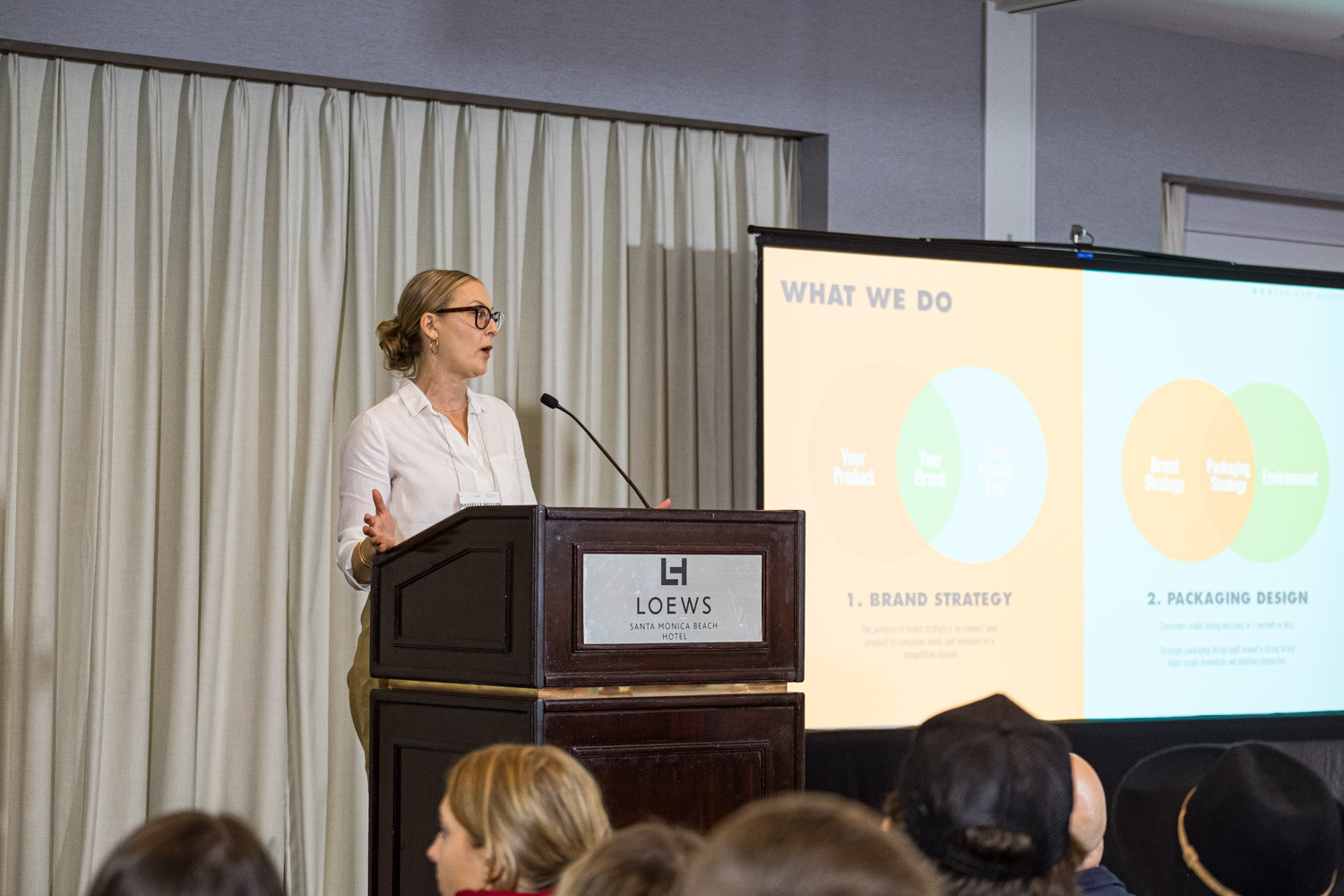 A woman in a white shirt stands at a Loews podium next to a screen titled What we do showing brand strategy and packaging design.
