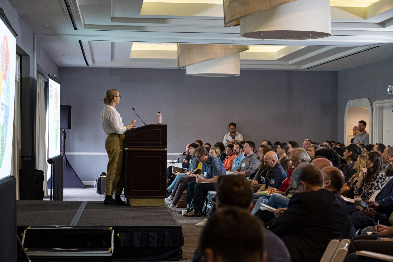 A woman at a podium on a low stage faces rows of seated attendees in a hotel meeting room.