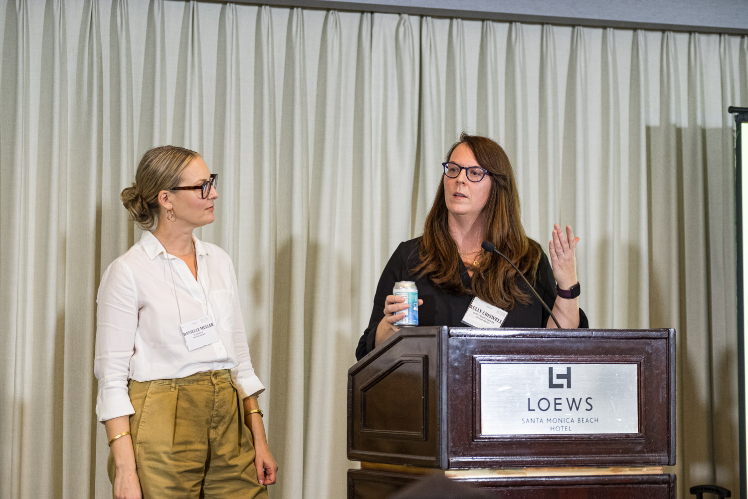 Two women stand at a Loews podium, one in a white shirt and one in a black top holding a drink can.