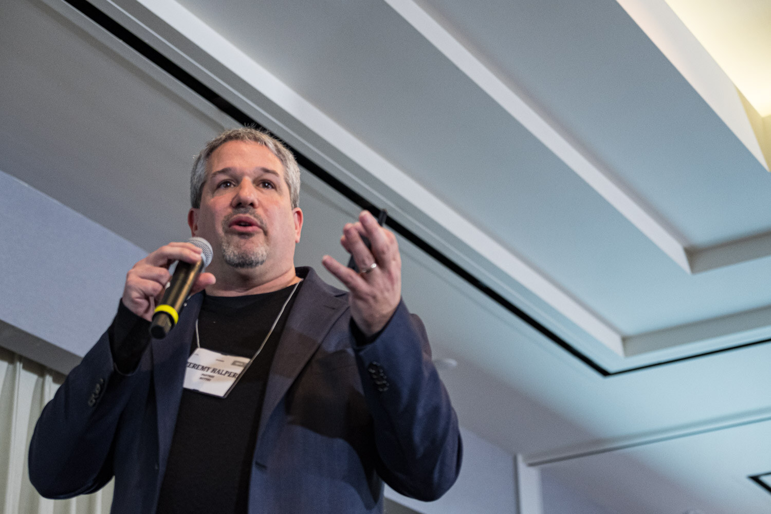 A man in a navy blazer and black shirt speaks into a handheld microphone and gestures with both hands.