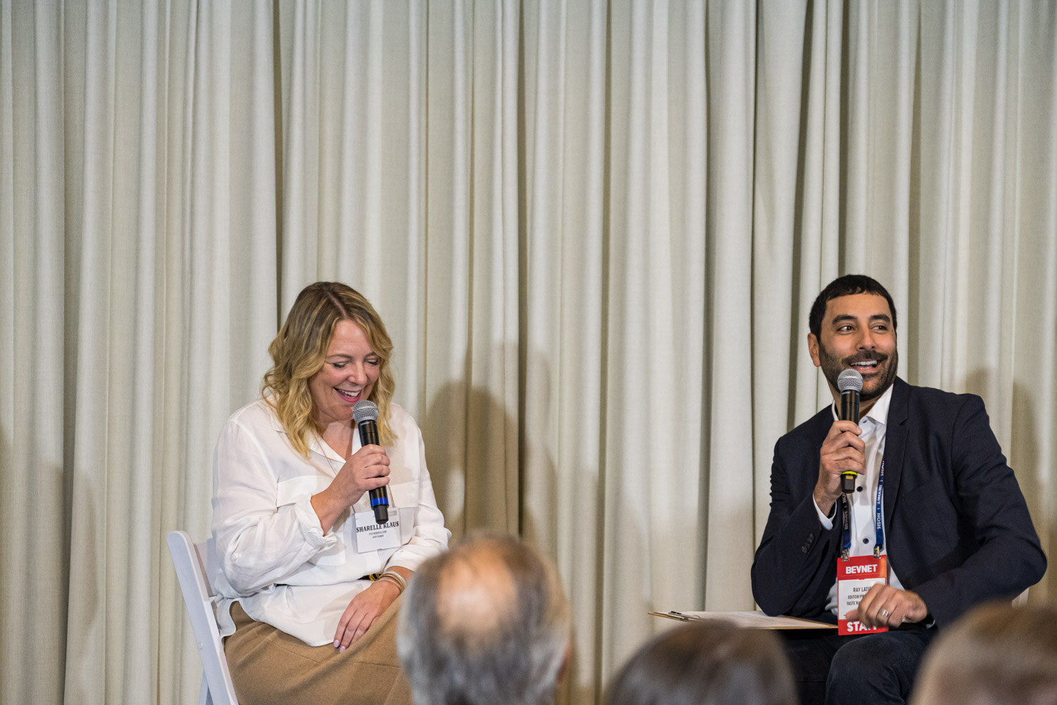 A woman in a white blouse laughs while holding a microphone next to a man in a navy blazer with a clipboard.