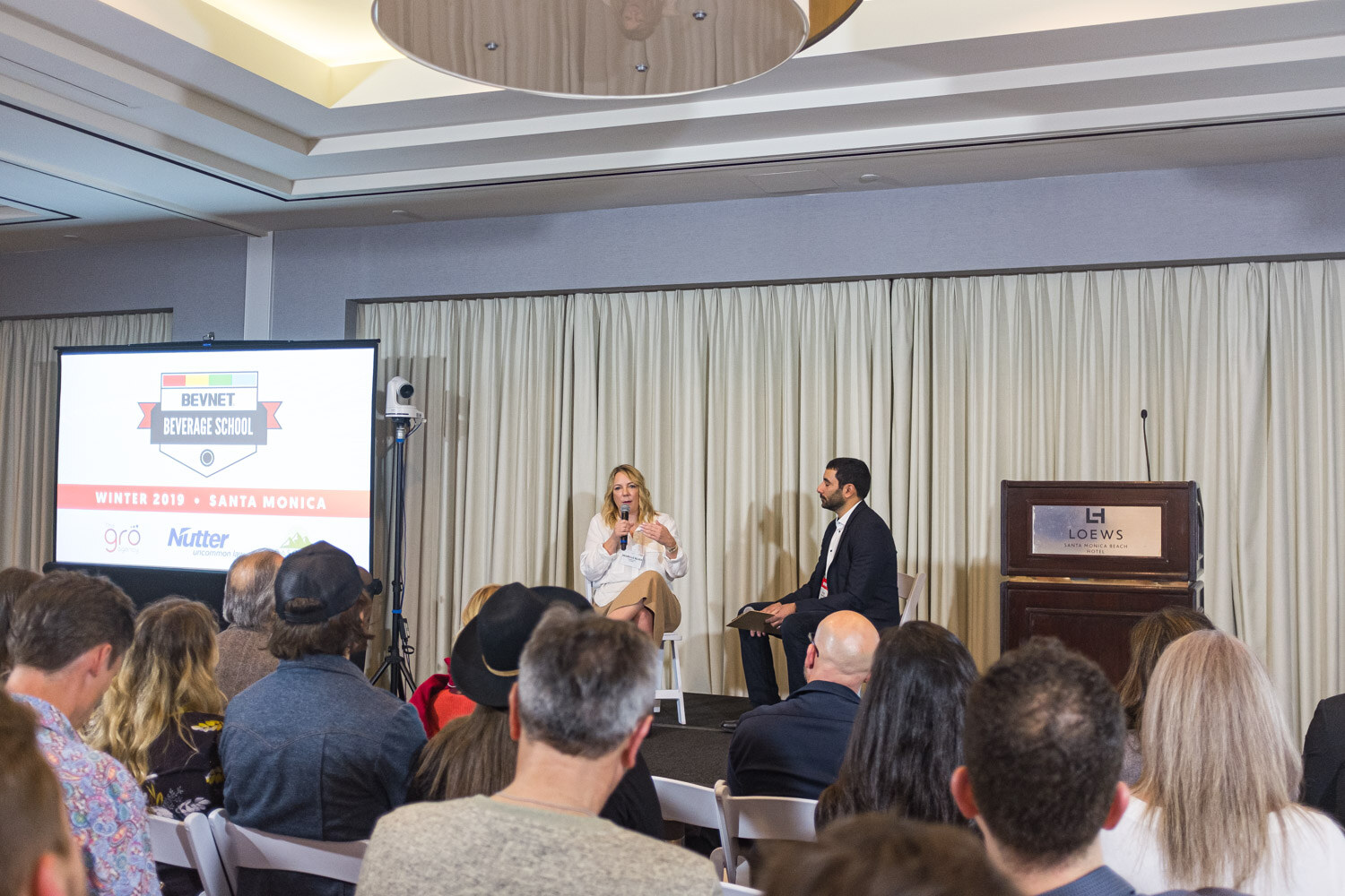 A woman with a microphone and a man with a clipboard sit on stage before an audience and a screen reading BevNET Beverage School Winter 2019 Santa Monica.