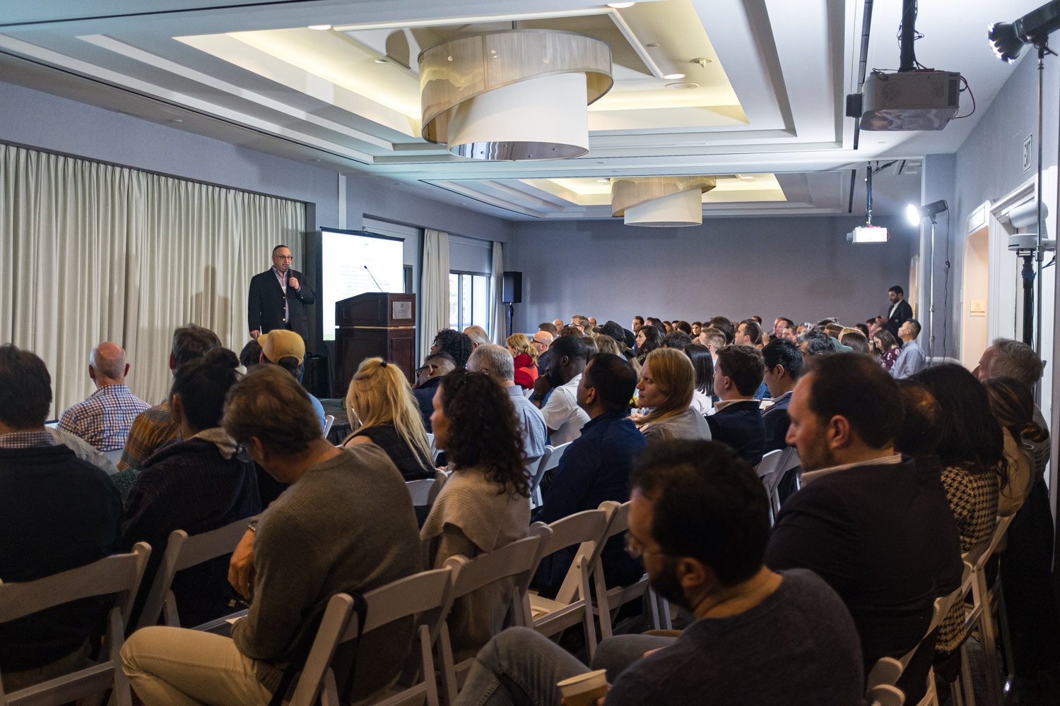 A packed meeting room of seated attendees facing a presenter at a wooden podium at the far end.
