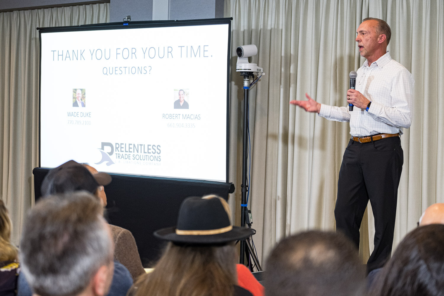 A man in a white shirt gestures beside a projection screen reading Thank you for your time, questions? with Relentless Trade Solutions branding.