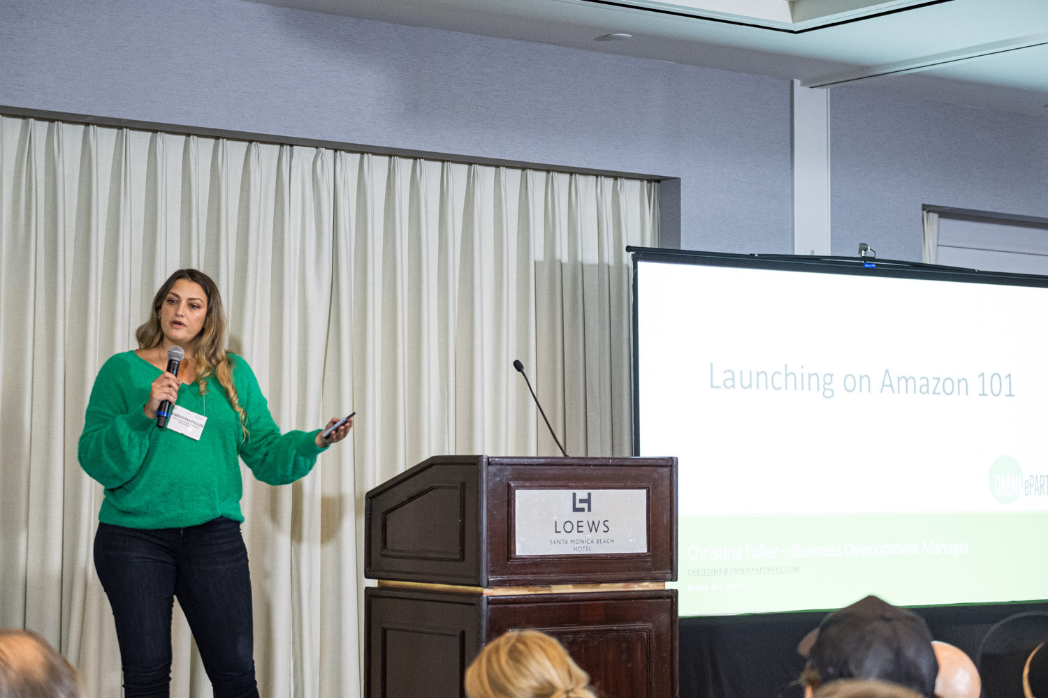 A woman in a green sweater holds a microphone next to a Loews podium and a projection screen reading Launching on Amazon 101.