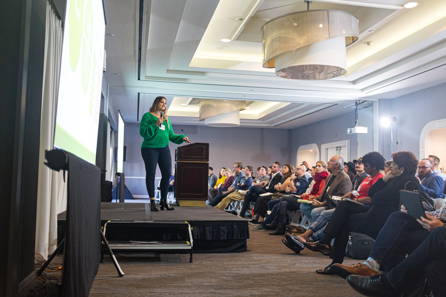 A woman in a green sweater stands on a low stage beside a podium while rows of attendees watch.