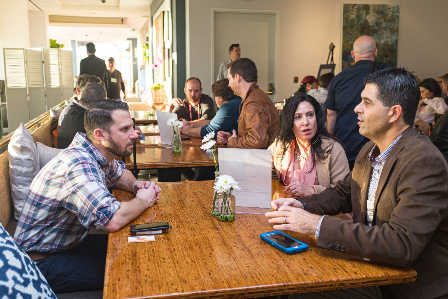 Five people sit around a wooden table with a table card and a small vase of white daisies.