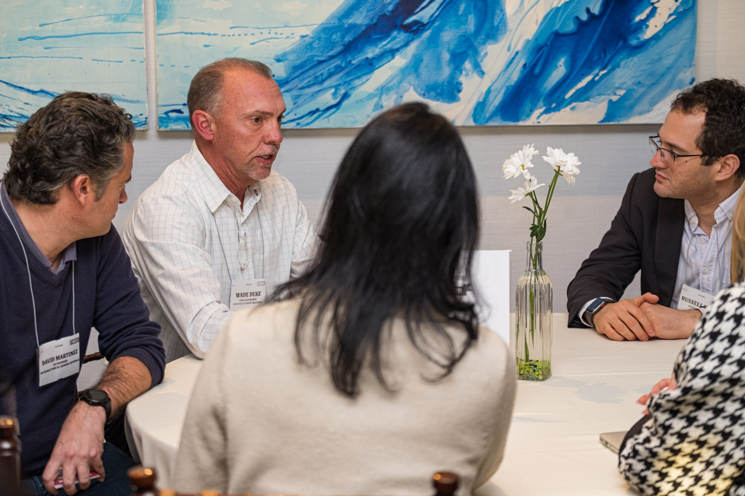A man in a white shirt talks with attendees at a white-clothed table with a vase of daisies.