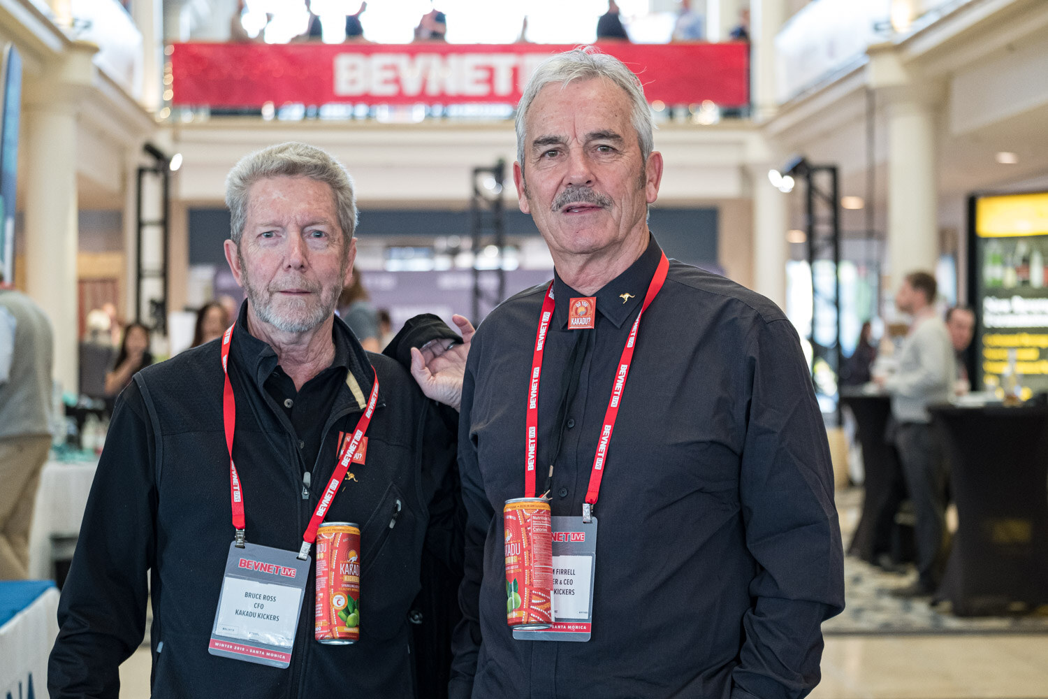 Two older men in black shirts with red lanyards pose in the atrium with a BevNET Live banner above them.