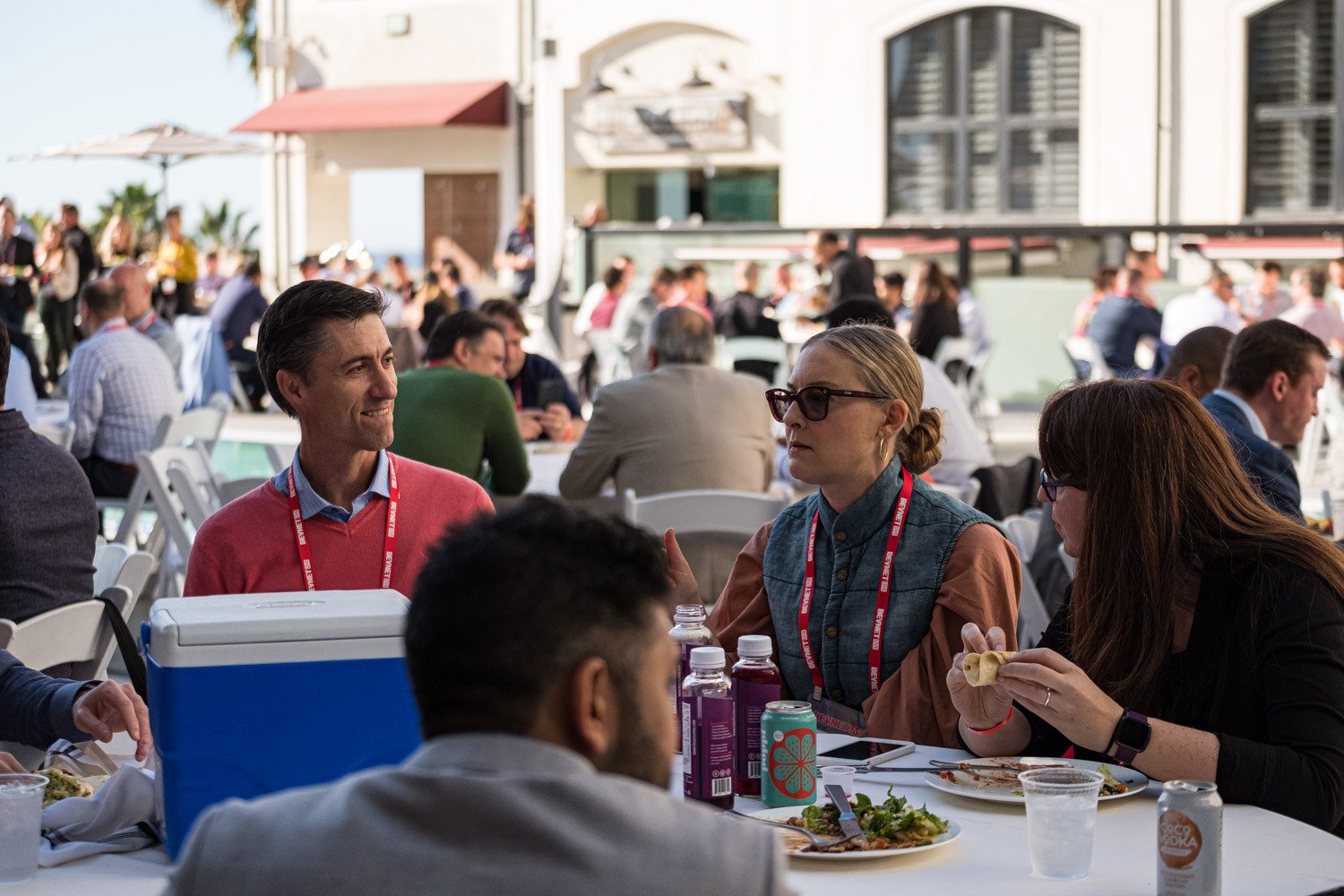 Attendees with red lanyards sit at round tables on a hotel terrace with a cooler and drinks.