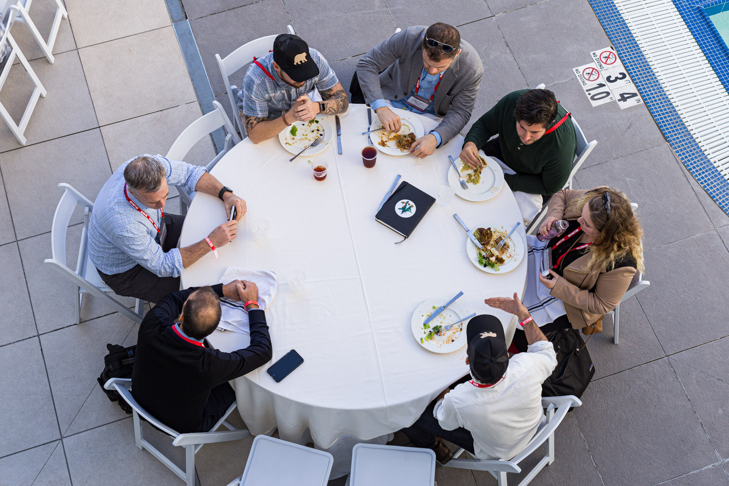 View from above of eight attendees seated around a round white table with plates and drinks next to the pool.