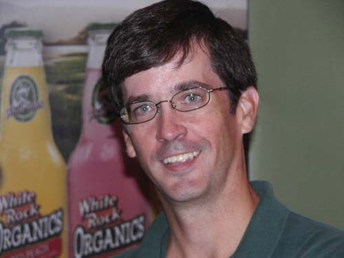 Close-up of a smiling man with dark hair and wire-framed glasses in a green shirt, in front of White Rock Organics bottles in yellow and pink.
