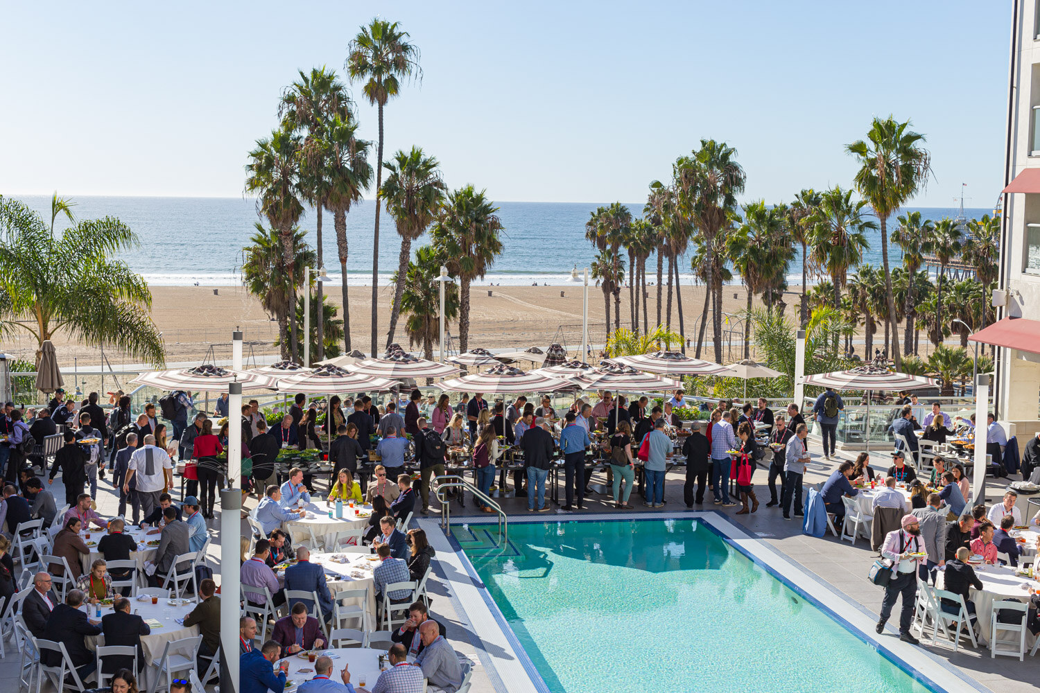 Wide view of a hotel pool terrace filled with attendees at tables under umbrellas, with palm trees and the ocean in the distance.