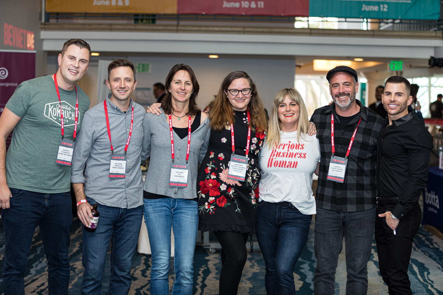 Seven people with conference lanyards stand together smiling in the hotel atrium, one wearing a Serious Business Woman shirt.