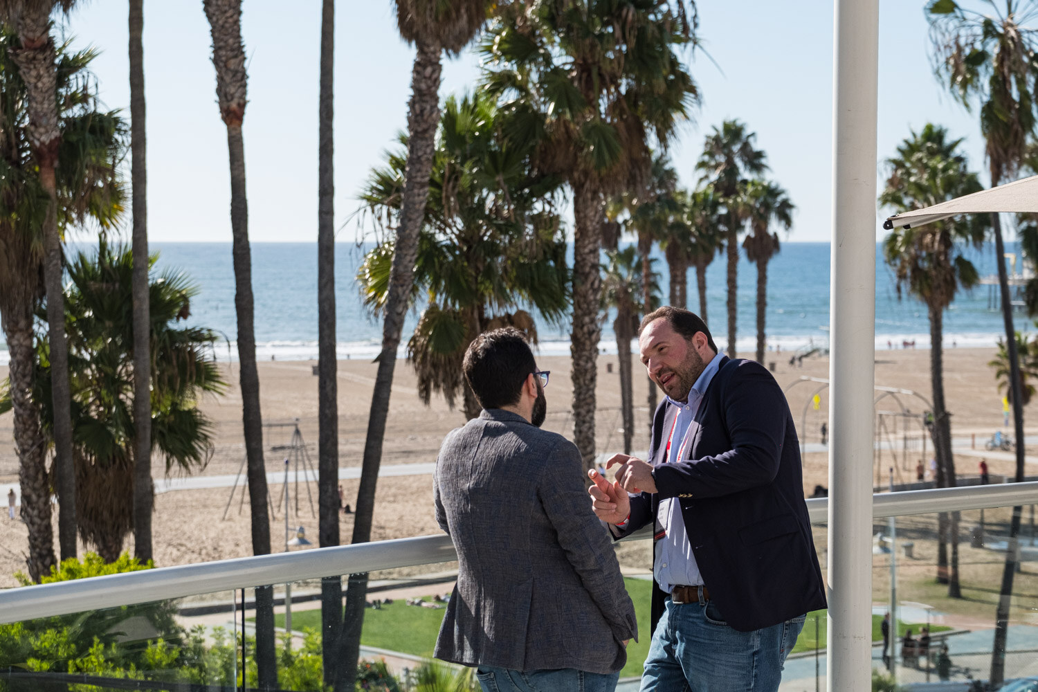Two men in blazers stand on a hotel balcony with palm trees, the beach and the ocean behind them.