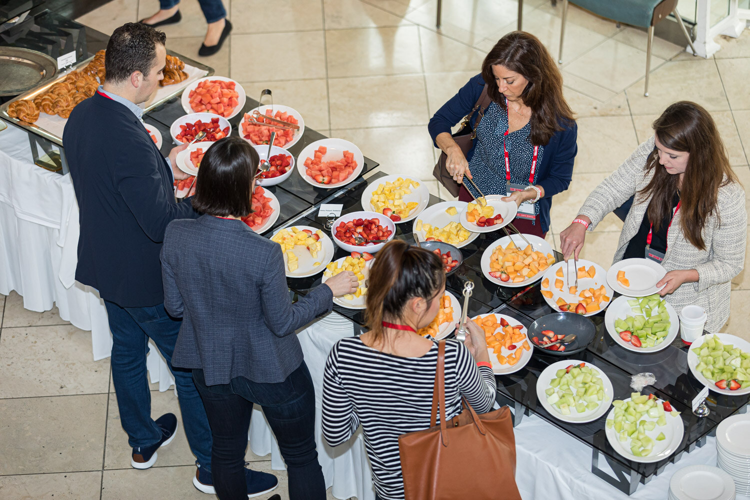 Overhead view of several attendees serving fruit from a buffet table with bowls of melon, strawberries and oranges.